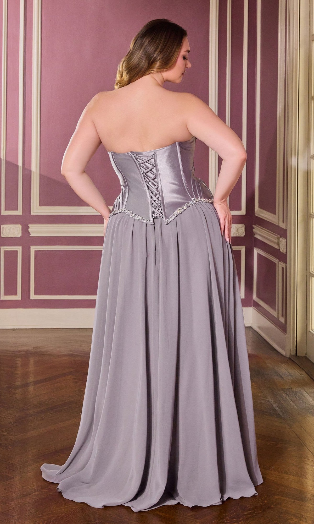 A woman in the Ladivine CD668C Long Plus-Size Prom Dress, a strapless silver-gray gown with a corset bodice, stands with her back to the camera in a room with pink and cream paneled walls and a wooden floor.